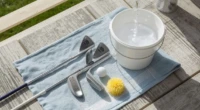A set of golf clubs with a bucket of soapy water, brush, and microfiber towel, ready for cleaning on a bright outdoor patio.